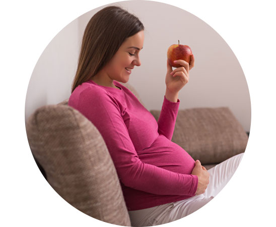 Image shows pregnant woman sitting on the sofa eating an apple